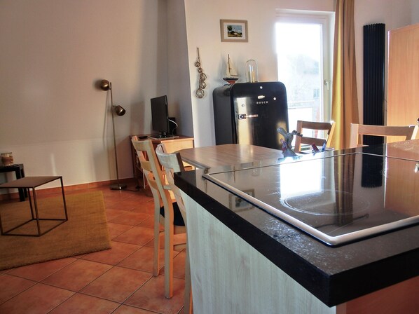 A modern kitchen with a black countertop, wooden chairs, a refrigerator, and a window with curtains.