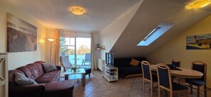 Ein gemütliches Wohnzimmer mit einer braunen Ledersofa, einem Holztisch und einem Essbereich mit Tisch und Stühlen. Ein Fernseher steht auf einem Schrank, ein Gemälde hängt an der Wand und über dem Essbereich gibt es ein Oberlicht.