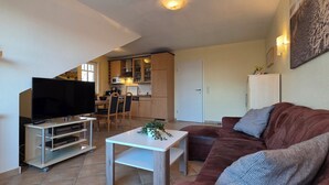 Un salon avec un téléviseur, un canapé et une petite table. Au fond, on aperçoit une cuisine équipée de placards et d’un four à micro-ondes.