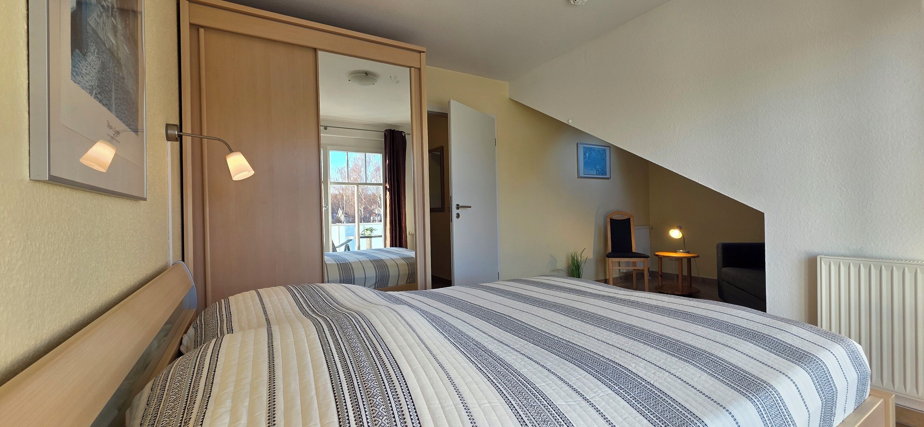A bedroom with a bed, a door leading to another room, a small table with a lamp, and a radiator.