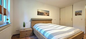 Een slaapkamer met een bed, een klein nachtkastje met een plant, een raam met jaloezieën en een deur.