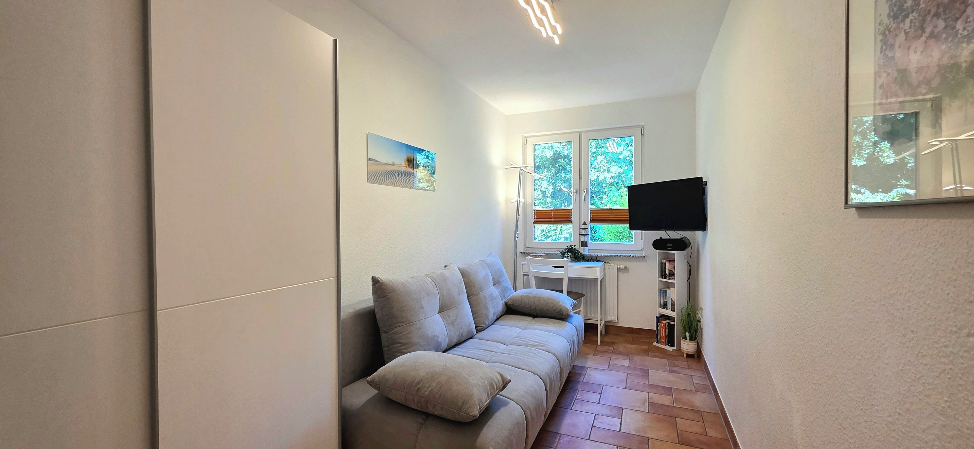 A narrow hallway with a sofa, a desk, a chair, a TV, and a window with blinds.