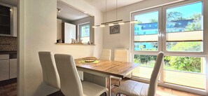 Een eetgedeelte met een houten tafel, witte stoelen, een spiegel en uitzicht op groen.
