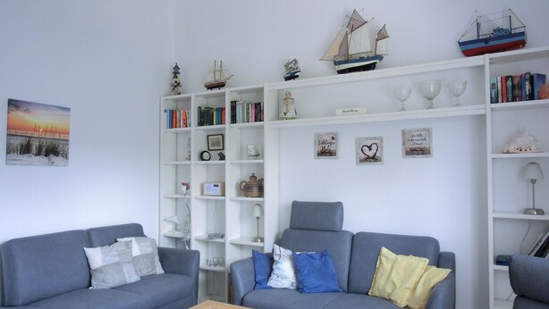 Een woonkamer met twee grijze banken, een glazen salontafel, een boekenkast vol boeken en decoratieve objecten, en een wand met ingekaderde foto's en een schilderij.