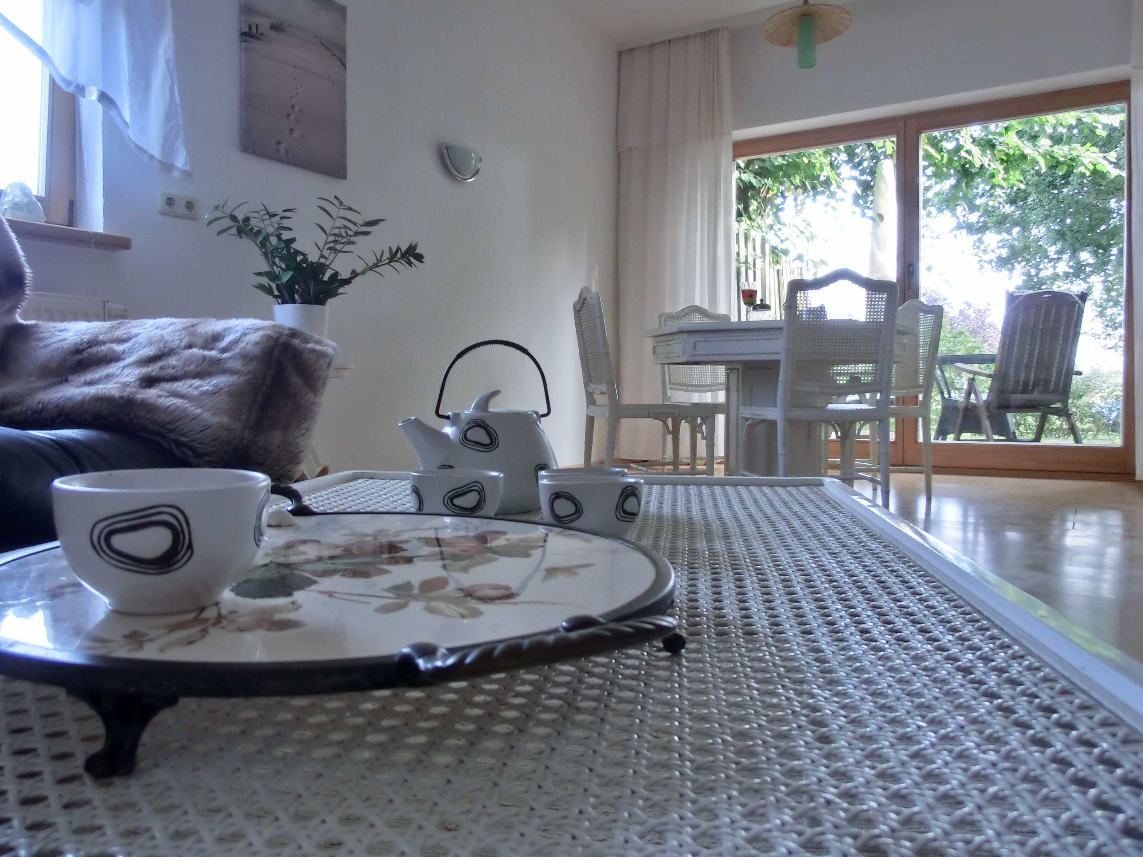 A cozy living room with a sofa, a table with a teapot and cups, a potted plant, and a view of the outdoors through a glass door.