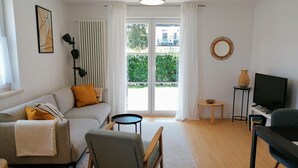 Un salon moderne avec un canapé, un fauteuil, une petite table, un téléviseur et une vue sur la verdure à l’extérieur.