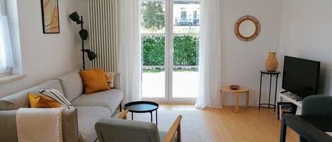A modern living room with a sofa, a chair, a small table, a TV, and a view of greenery outside.