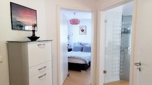 Un couloir menant à une chambre à coucher, où se trouvent un lit, une commode et un tableau accroché au mur.
