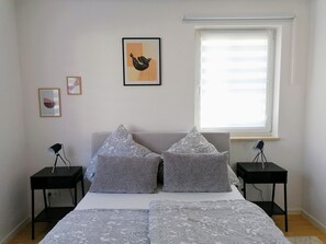 Un dormitorio con una cama, dos mesitas de noche, una ventana con persianas y dos cuadros enmarcados en la pared.