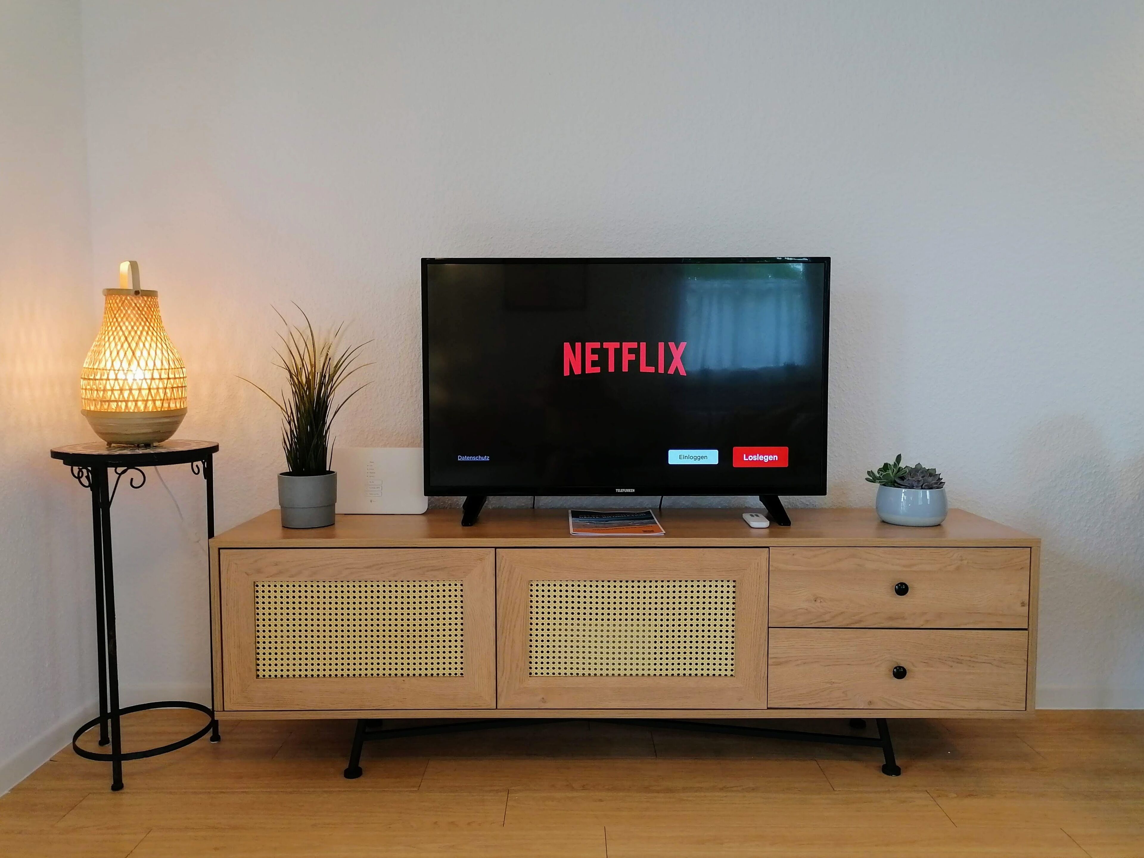 Ein Fernseher steht auf einer Holzkommode, wobei die Netflix-Oberfläche angezeigt wird. Dazu gehören eine Lampe und zwei Zimmerpflanzen.