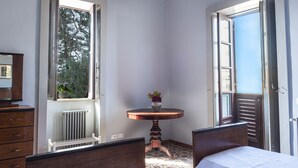 A bedroom with a bed, a dresser, a table with a vase, and a window with a view of trees. - BAGLIO OF SANTA MARINA (Milazzo)