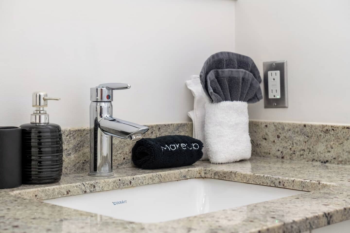 Un lavabo de salle de bain avec un robinet chromé, un distributeur de savon noir et une serviette ornée d’une broderie « MOUSUP ».