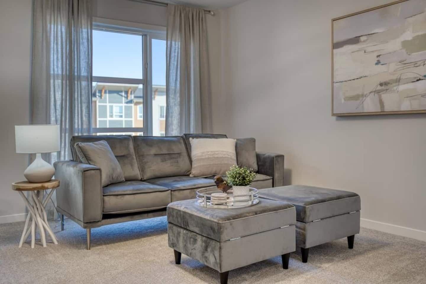 Ein modernes Wohnzimmer mit einem grauen Sofa, einem passenden Hocker, einem Beistelltisch mit Lampe und einem gerahmten abstrakten Gemälde an der Wand.