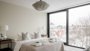 Un dormitorio moderno con un ventanal grande, una cama con colcha color beige, dos mesitas de noche con lámparas, y vistas a un paisaje urbano nevado.