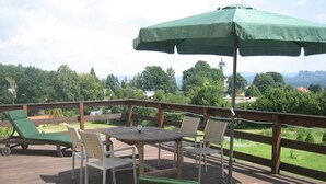 A wooden deck with a table and chairs, a green umbrella, and a view of trees and buildings. - FeWo Zirkelstein, Landhaus Böhmer***** (Papstdorf)