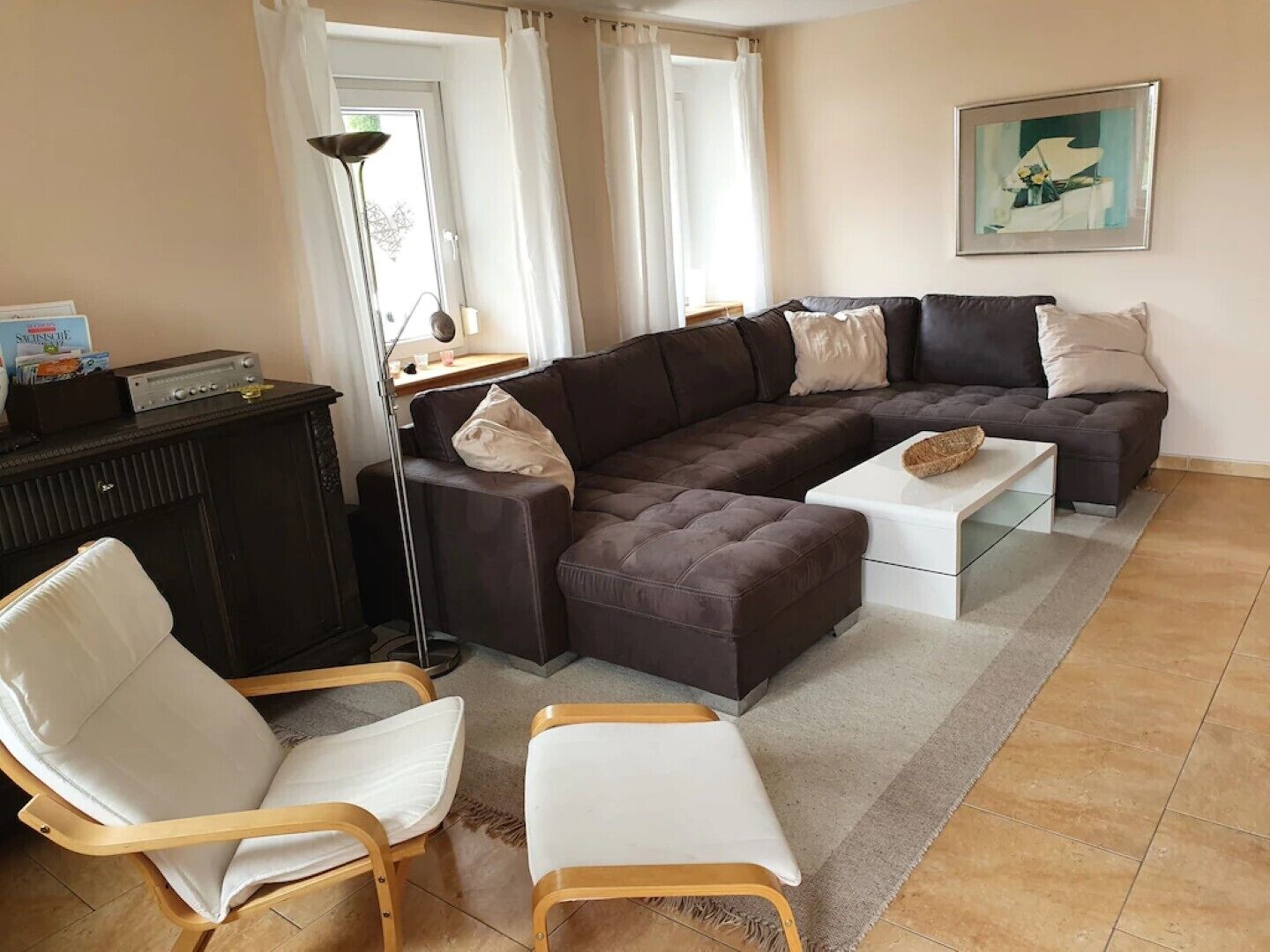 Ein Wohnzimmer mit einem Ecksofa, zwei weißen Sessel, einem Couchtisch, einer dunklen Kommode und einer Stehlampe.