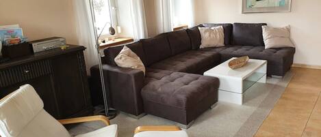 Ein Wohnzimmer mit einem Ecksofa, zwei weißen Sessel, einem Couchtisch, einer dunklen Kommode und einer Stehlampe.