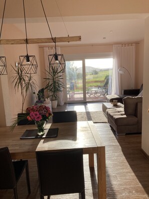 Un coin repas avec une table en bois, des chaises et un vase de fleurs. Le salon comprend un canapé et une lampe.