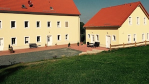 Ein Gebäude mit rotem Ziegeldach und gelber Fassade, mit mehreren Fenstern und einer kleinen Außenbestuhlung.