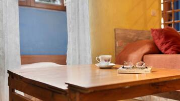 Una mesa de madera con una taza y vasos, un libro y un cojín sobre un sofá en una habitación con pared azul y una ventana.