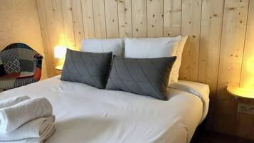 A neatly made bed with white linens and two grey pillows in a room with wooden panel walls and a small round table with a lamp.