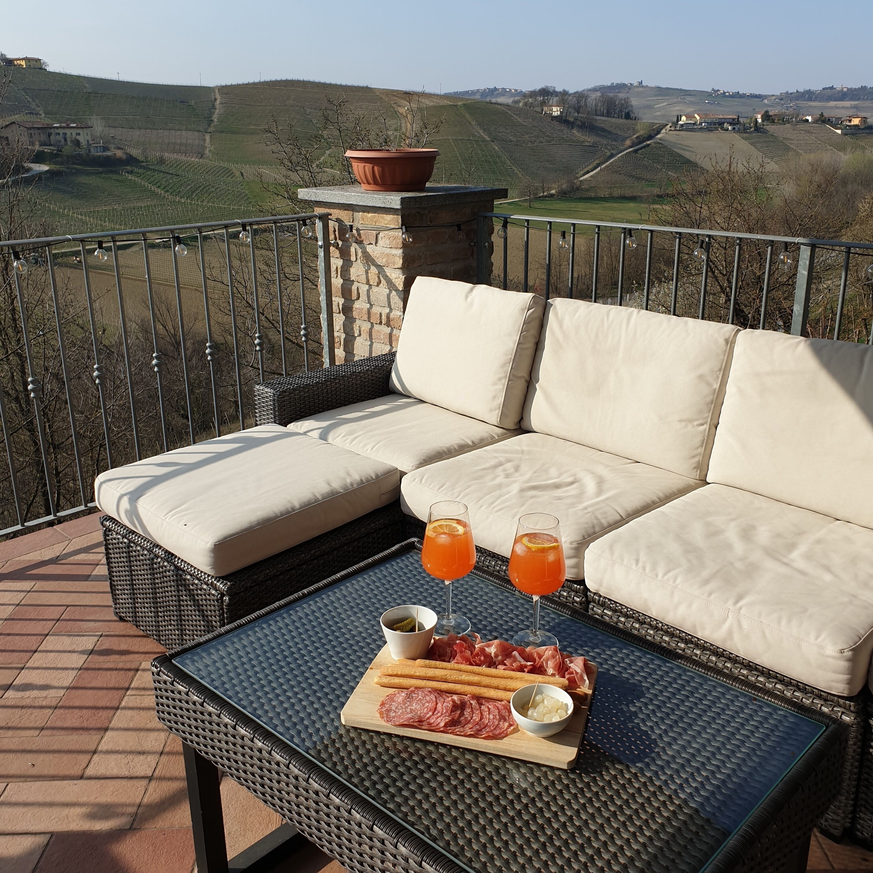 A balcony with a wicker sofa, a table with drinks and charcuterie, and a view of terraced fields.