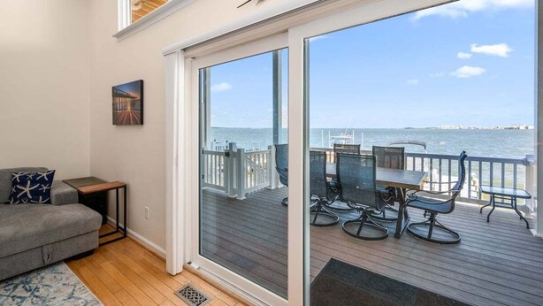 A balcony with a view of the sea, a dining area with chairs and a table, a gray sofa, and a framed picture on the wall.