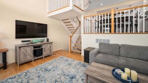 A spacious living room with a staircase, a flat-screen TV, a grey sofa, and a wooden coffee table.