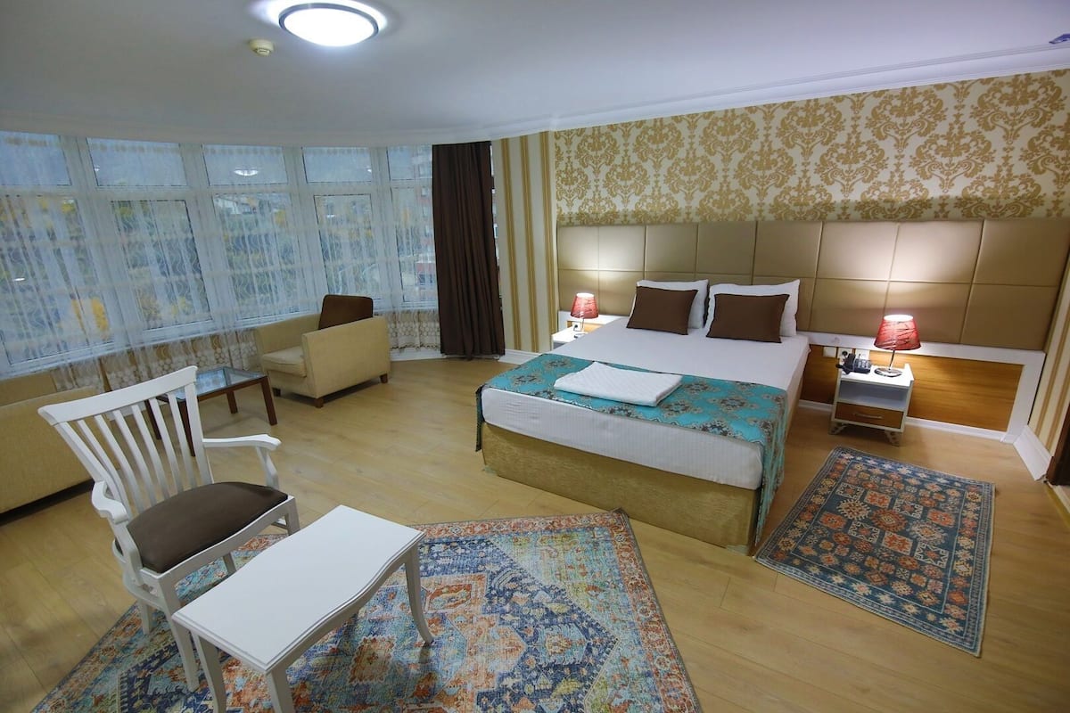 A hotel room with a large bed, a desk, a chair, a small table, and a nightstand with a lamp.