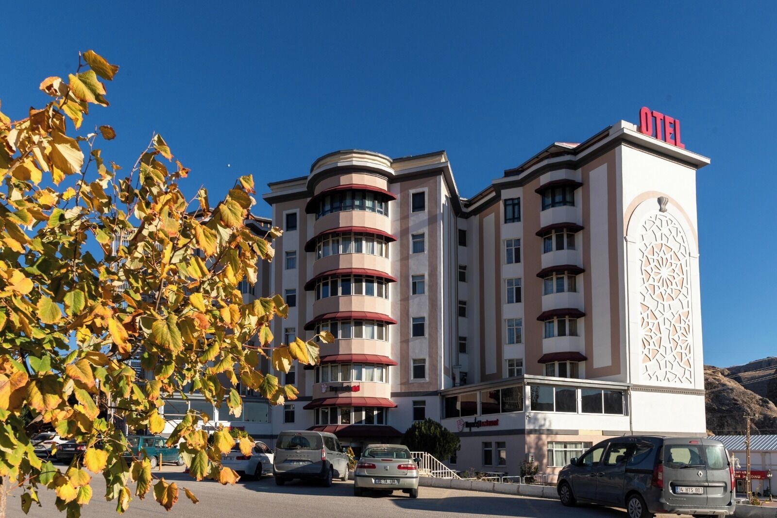 A multi-story hotel with a prominent "OTEL" sign, featuring a modern architectural design with balconies and a parking area in front.
