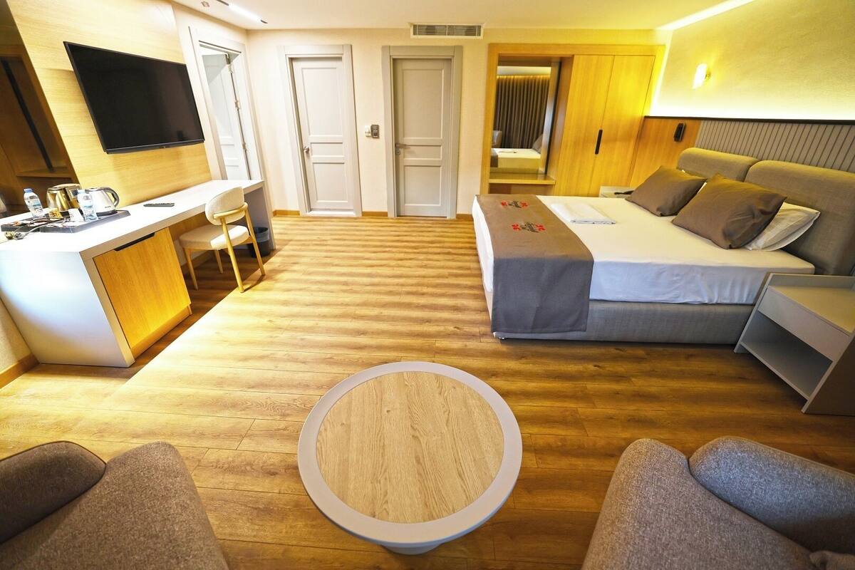 A hotel room with a bed, a sofa, a round coffee table, a TV, and a desk with a kettle.