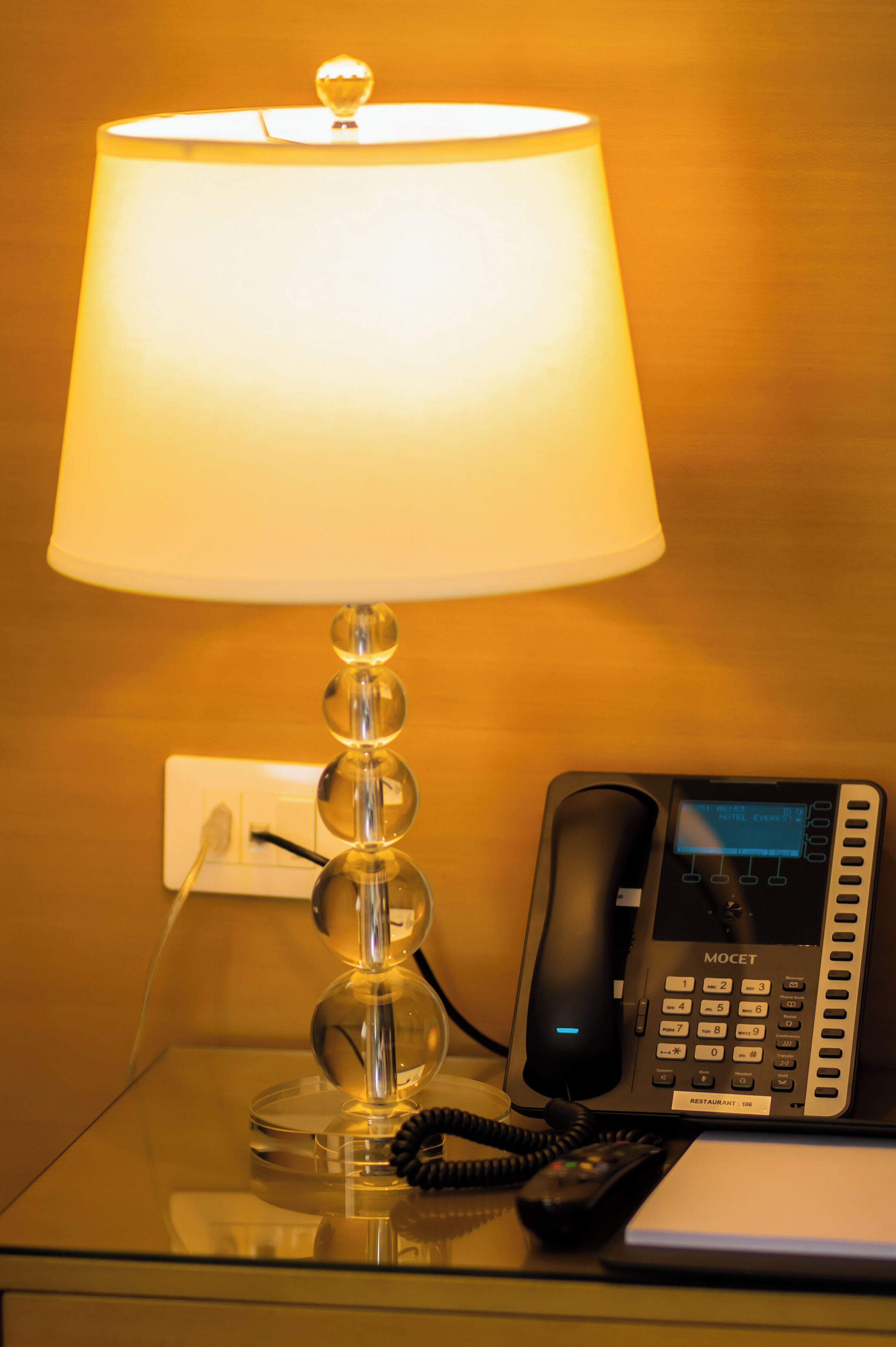 Een glazen tafel met een lamp, telefoon en notitieboekje.