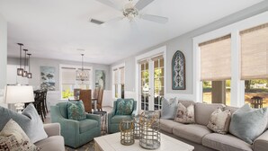 Un salon spacieux avec un canapé beige, un fauteuil turquoise et une table basse blanche. On y trouve des lanternes décoratives sur la table, un ventilateur de plafond et une grande fenêtre avec des stores.