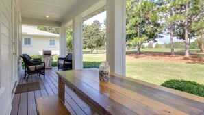 Une véranda couverte avec une table et des chaises en bois, un barbecue et une vue sur une cour verdoyante bordée d’arbres.