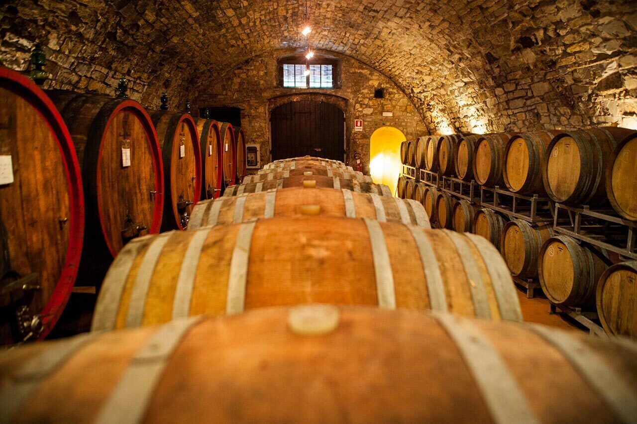 Una bodega con barricas de madera dispuestas en filas, techo de piedra y una puerta central.