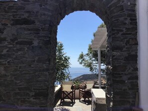 Un arco de piedra que da a un patio con mobiliario exterior y una pérgola, con vistas a un paisaje costero.