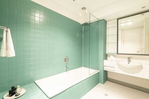 Um banheiro com azulejos verde-água, chuveiro com box de vidro, banheira branca e pia com bancada salpicada.