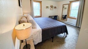 Un dormitorio con cama, escritorio, silla, lámpara y dos ventanas.