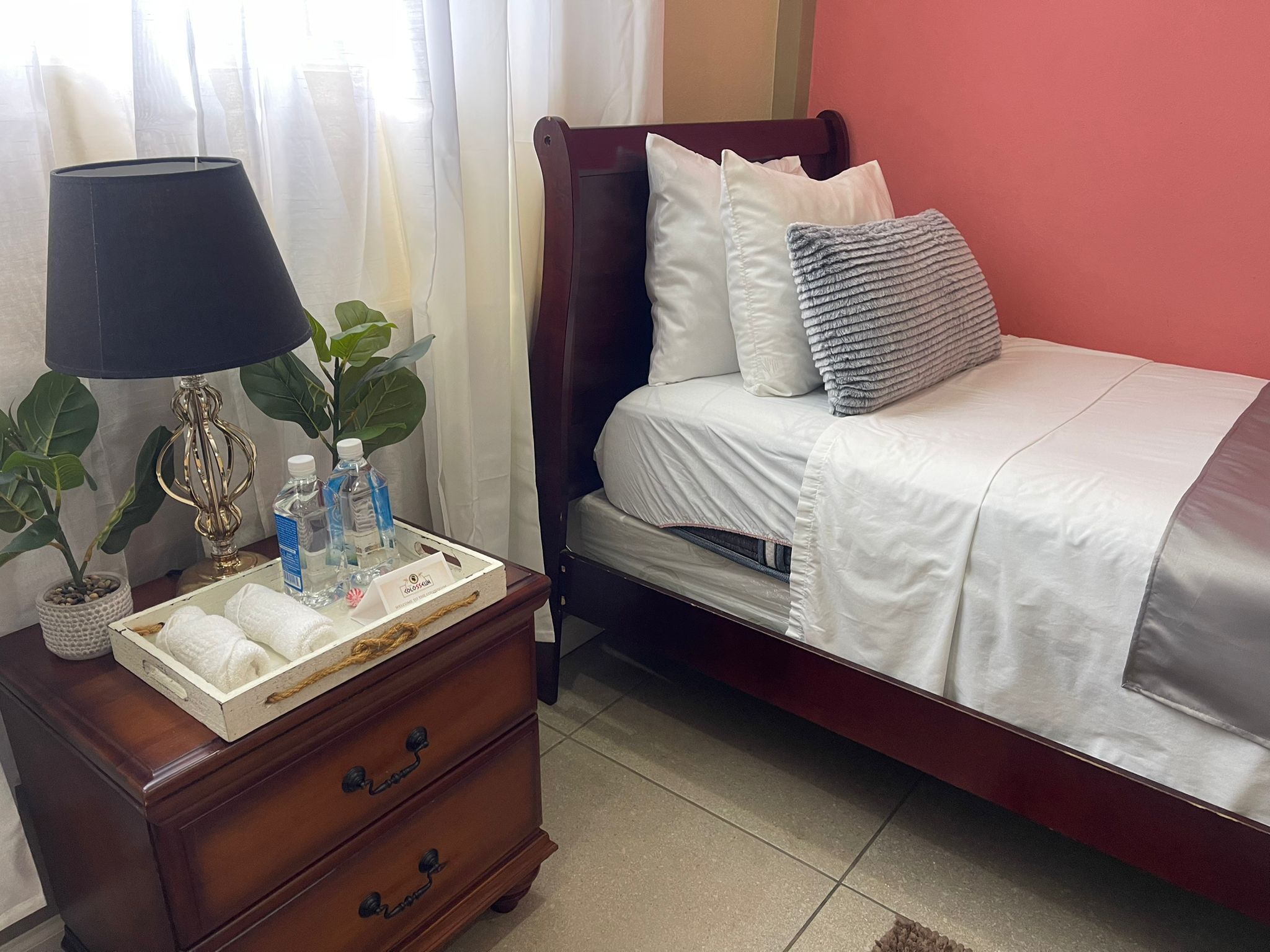 Un lit bien fait, avec des oreillers et une couverture, une table de chevet avec une lampe, une plante et un plateau contenant des articles de toilette.