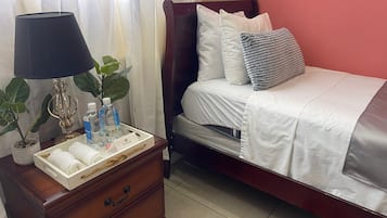 Una cama bien hecha con almohadas y una colcha, una mesita de noche con una lámpara, una planta y una bandeja con artículos de aseo.