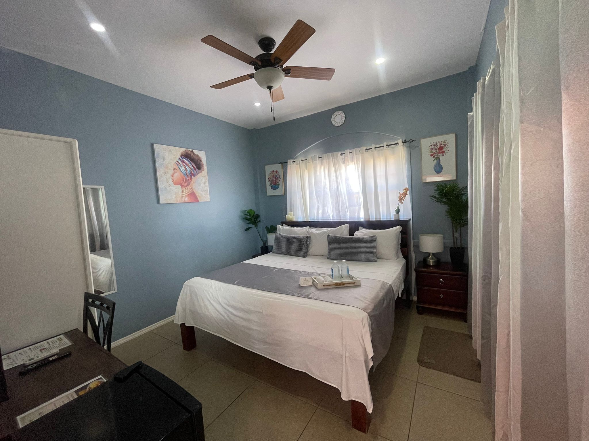 Un dormitorio con cama, ventilador de techo, mesita de noche, espejo y ventana con cortinas.