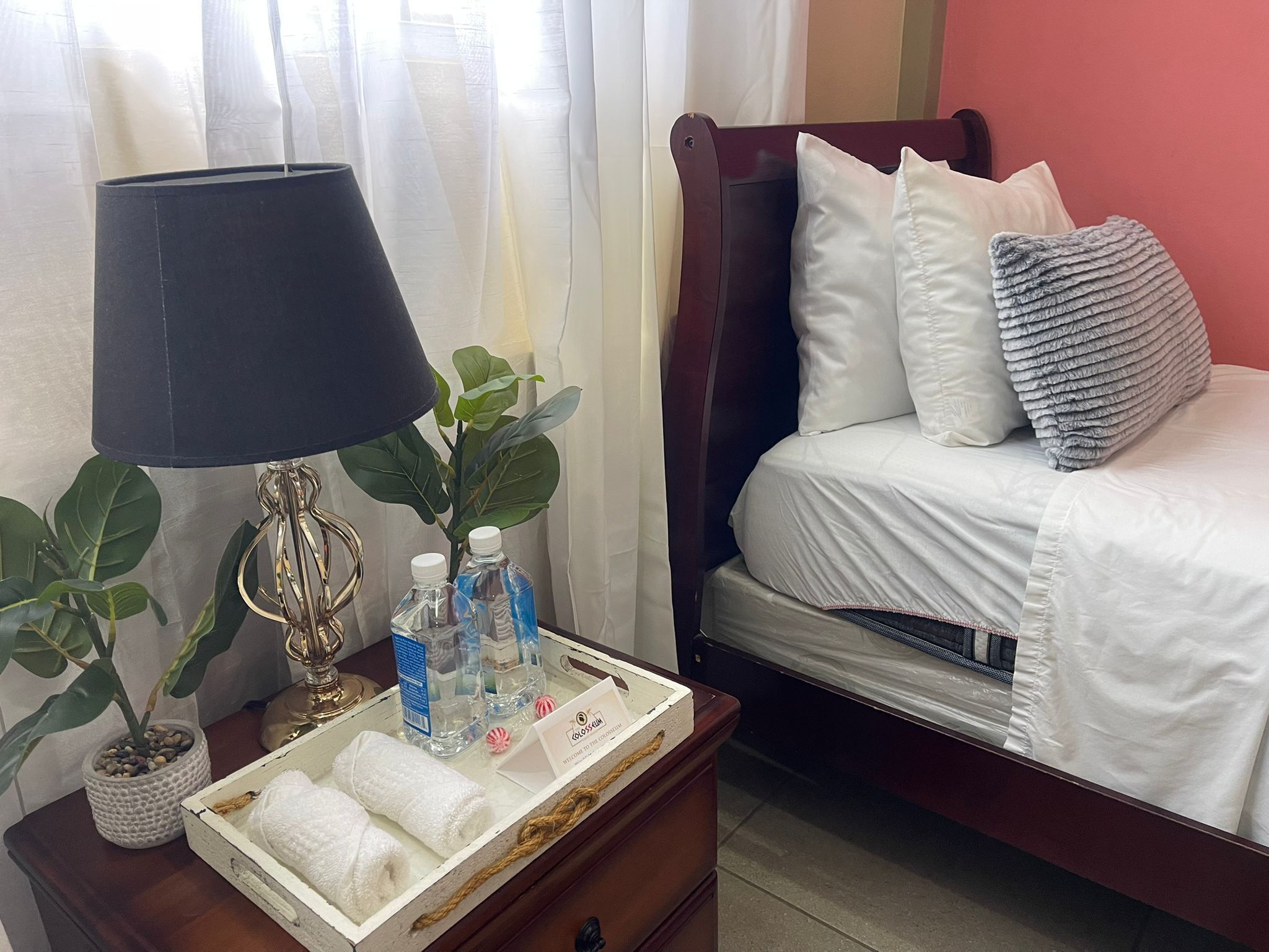 A neatly made bed with white linens, a bedside lamp, a small plant, and a tray with towels and toiletries.