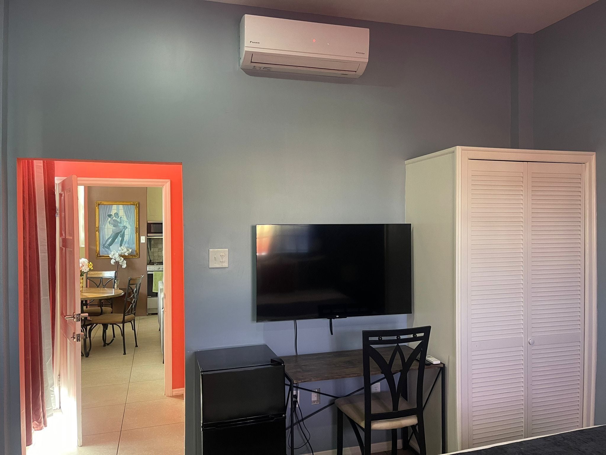 A room with a flat-screen TV mounted on the wall, a small desk with a chair, a white closet, and a red-framed doorway leading to another room.