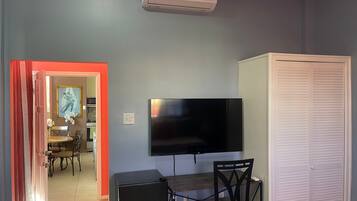 A room with a flat-screen TV mounted on the wall, a small desk with a chair, a white closet, and a red-framed doorway leading to another room.