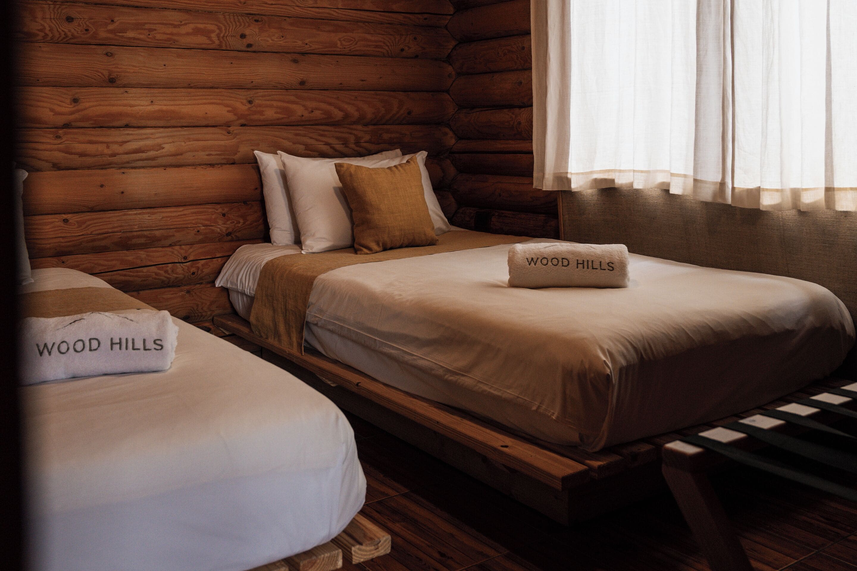 A cabin-style room with two beds, each with a 'WOOD HILLS' towel. The room has wooden walls and a window with curtains.