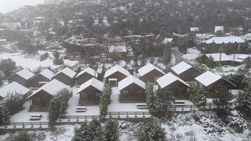 A snow-covered resort with multiple chalet-style cabins arranged in rows, surrounded by trees and a hilly backdrop.