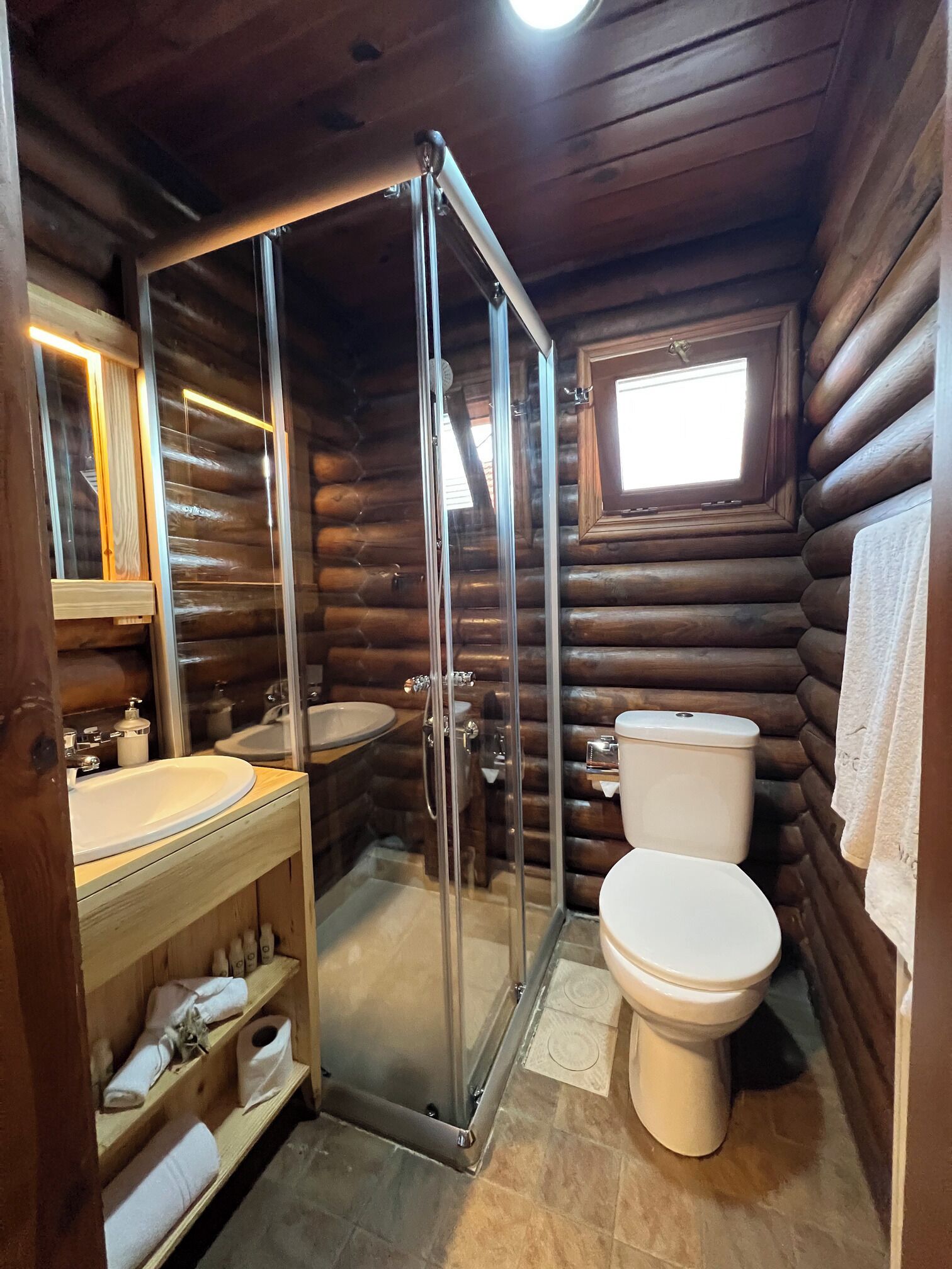 A log cabin bathroom with a glass-enclosed shower, a sink, a toilet, and a window.