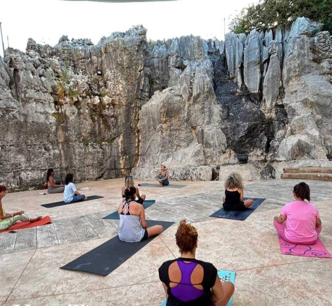 Eine Gruppe Menschen sitzt auf Yogamatten im Freien, im Hintergrund eine felsige Klippe.