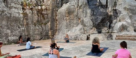 A group of people sitting on yoga mats outdoors with a rocky cliff in the background.