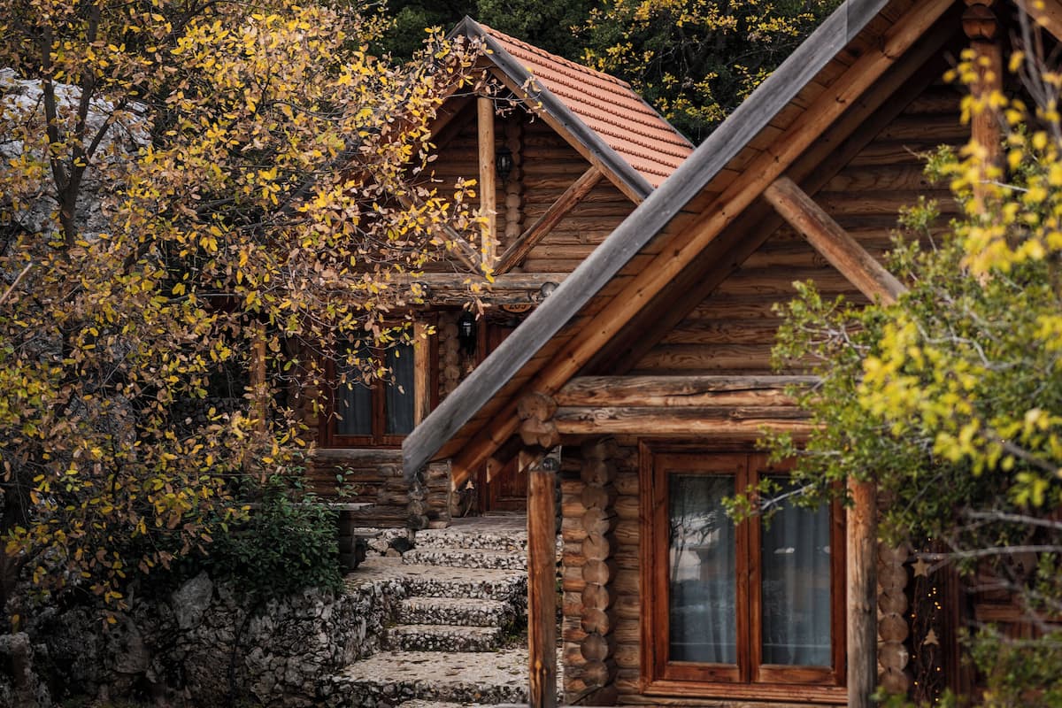 A rustic log cabin with a stone staircase leading to the entrance, surrounded by autumn foliage.